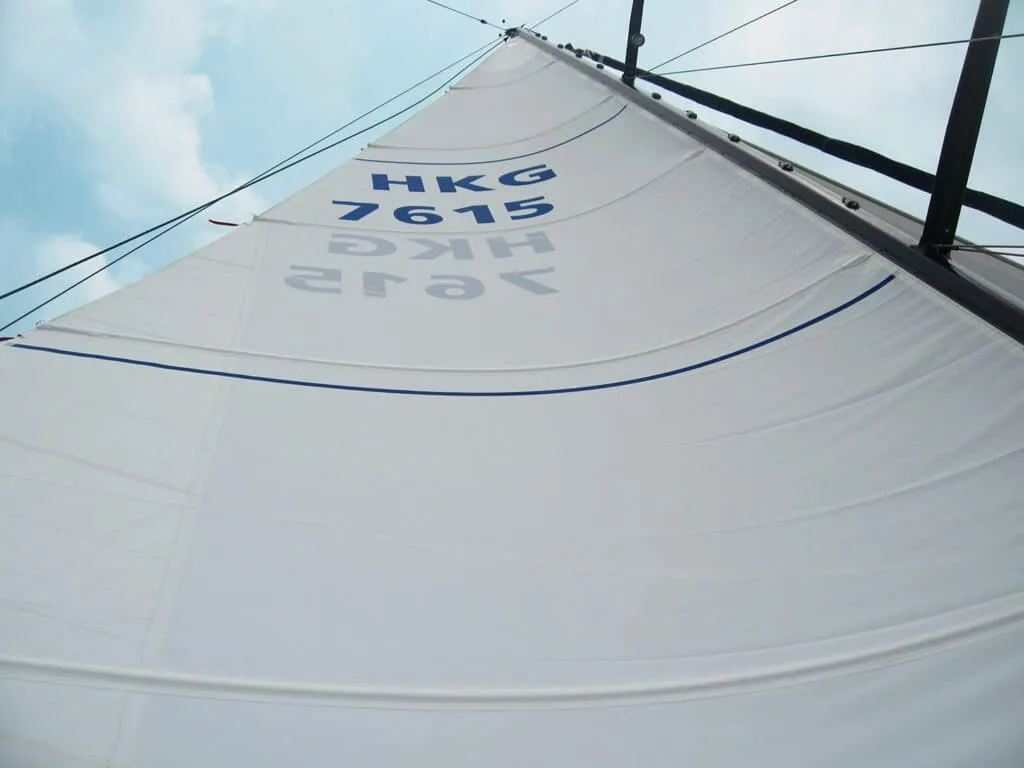 Upward view of a woven Dacron mainsail with sail number HKG 7615 against a hazy sky
