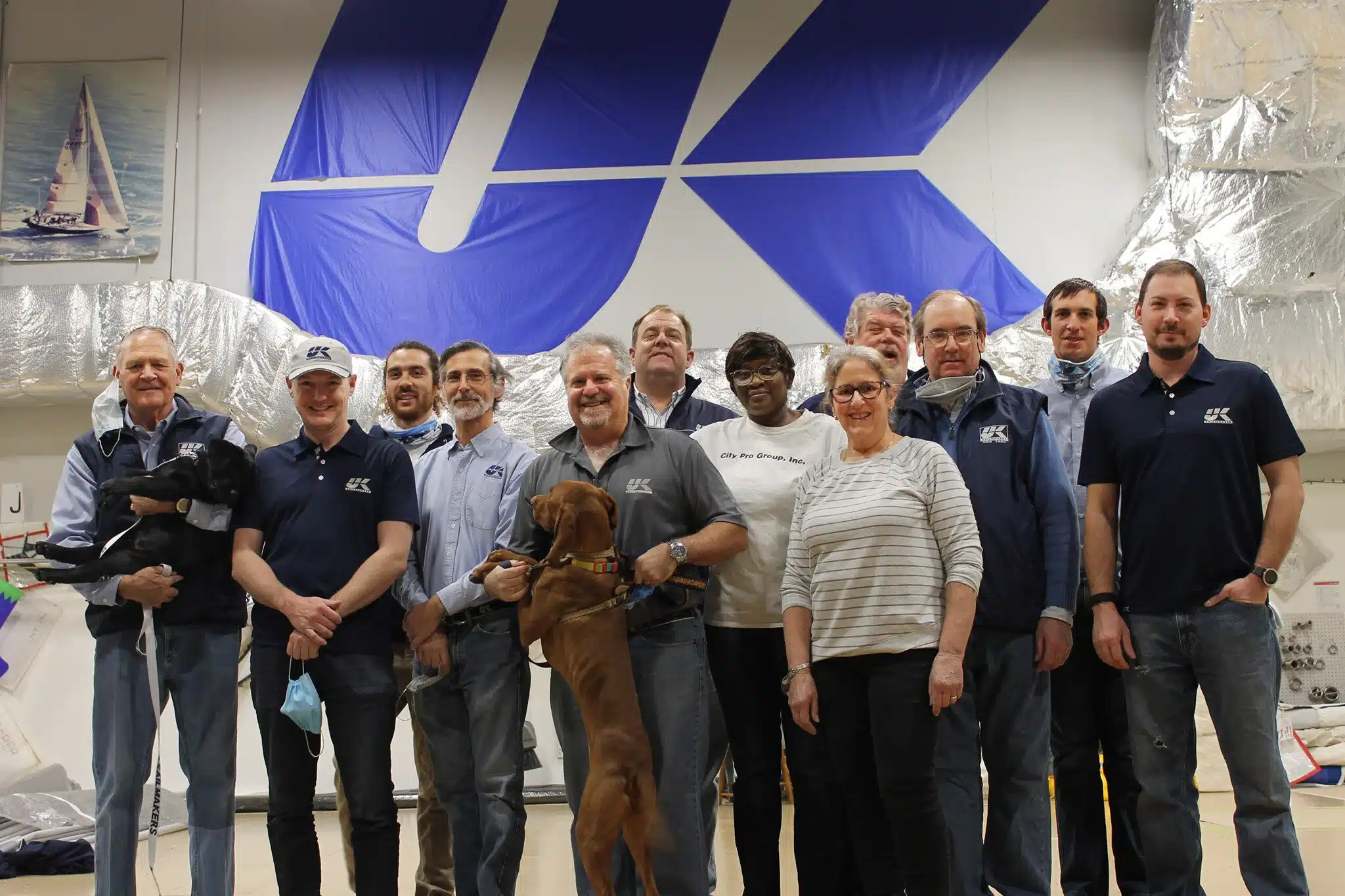 UK Sailmakers Northeast team posed in the loft in front of a large UK logo banner, with a dog