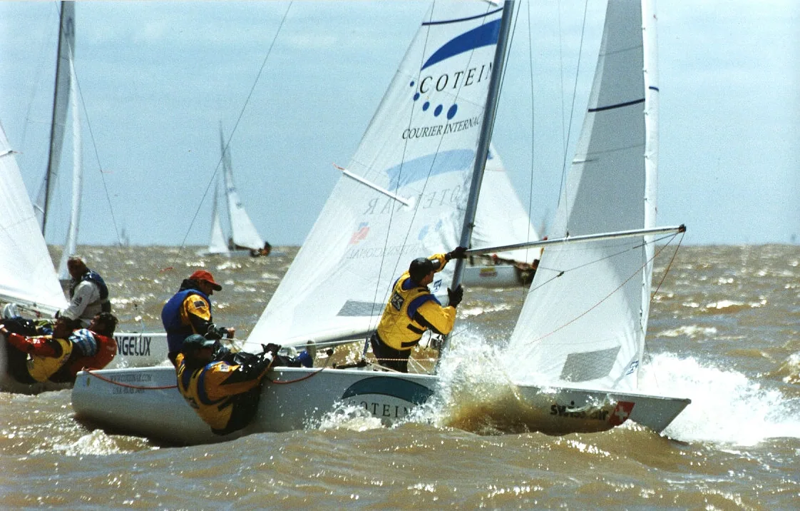 Two Solings racing hard with spray flying, one flying a sponsor-branded sail
