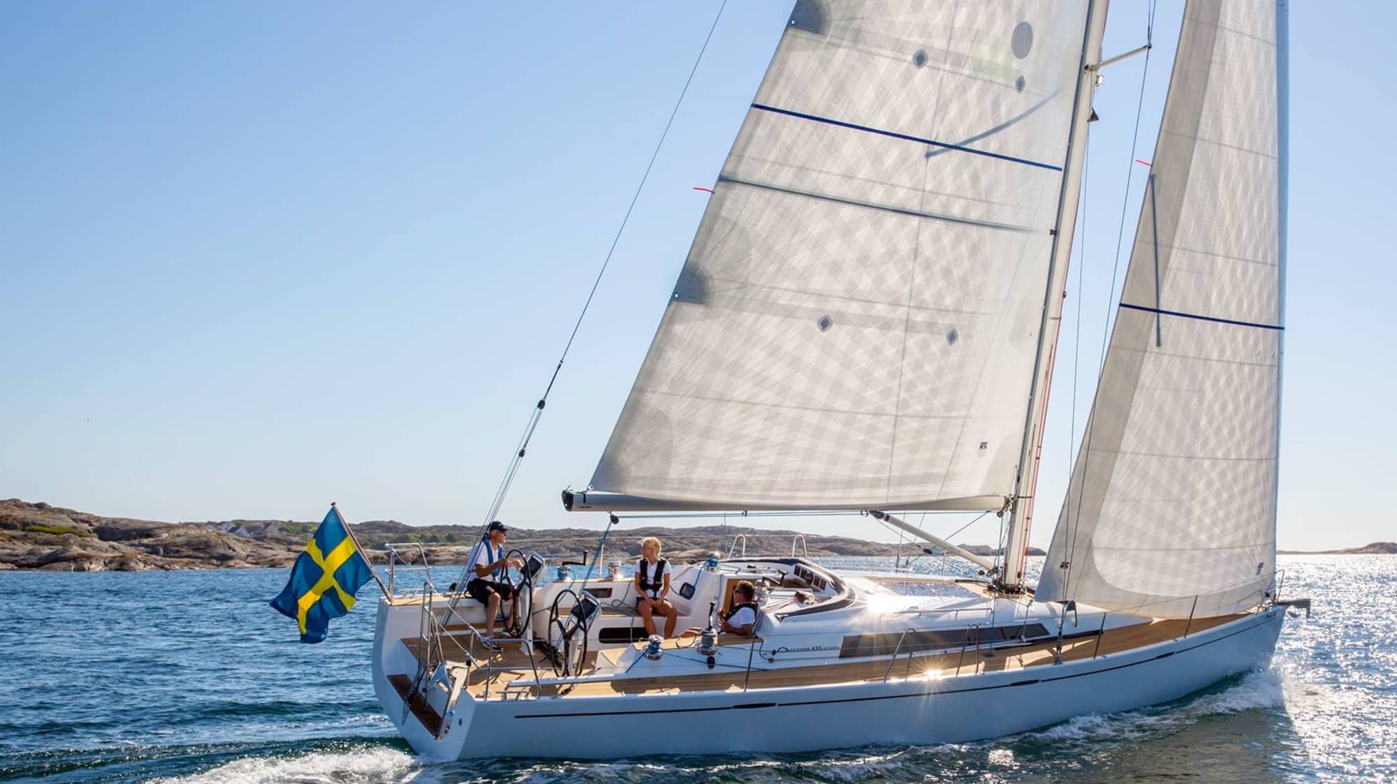 Cruising sloop with a Swedish courtesy flag sailing past a rocky coastline
