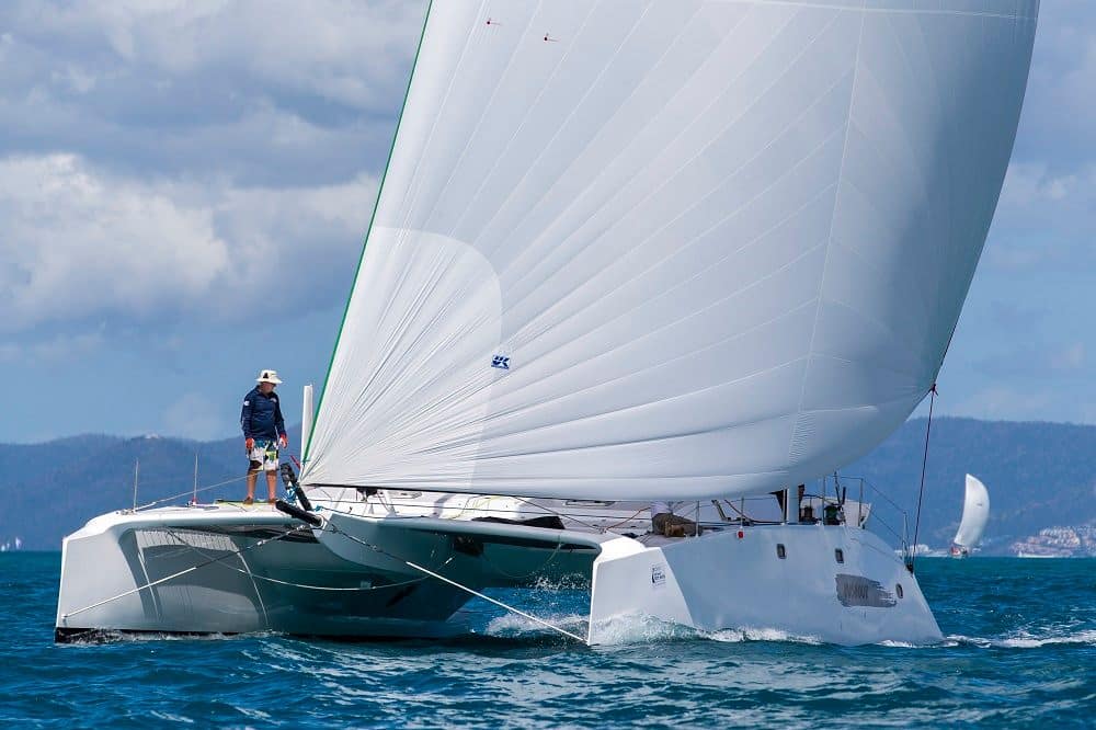 Trimaran with crew balanced on the ama, sailing near a coastal city skyline