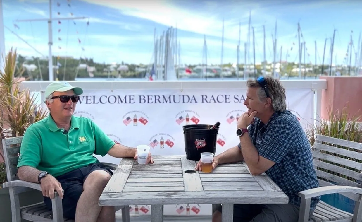 Two sailors relaxing with drinks under a 'Welcome Bermuda Race Sailors' banner
