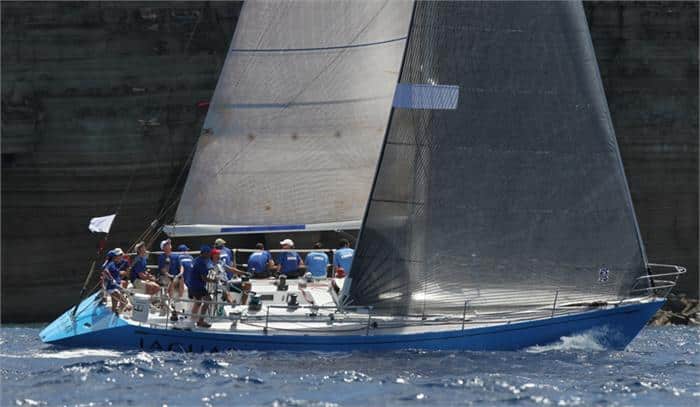 Racing sailboat heeled over with crew hiking on the rail near coastal cliffs