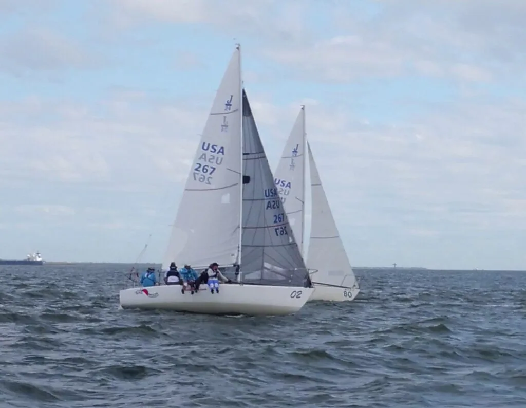 Two J/24s, sail number USA 267 leading, racing upwind near a container ship at J/Fest