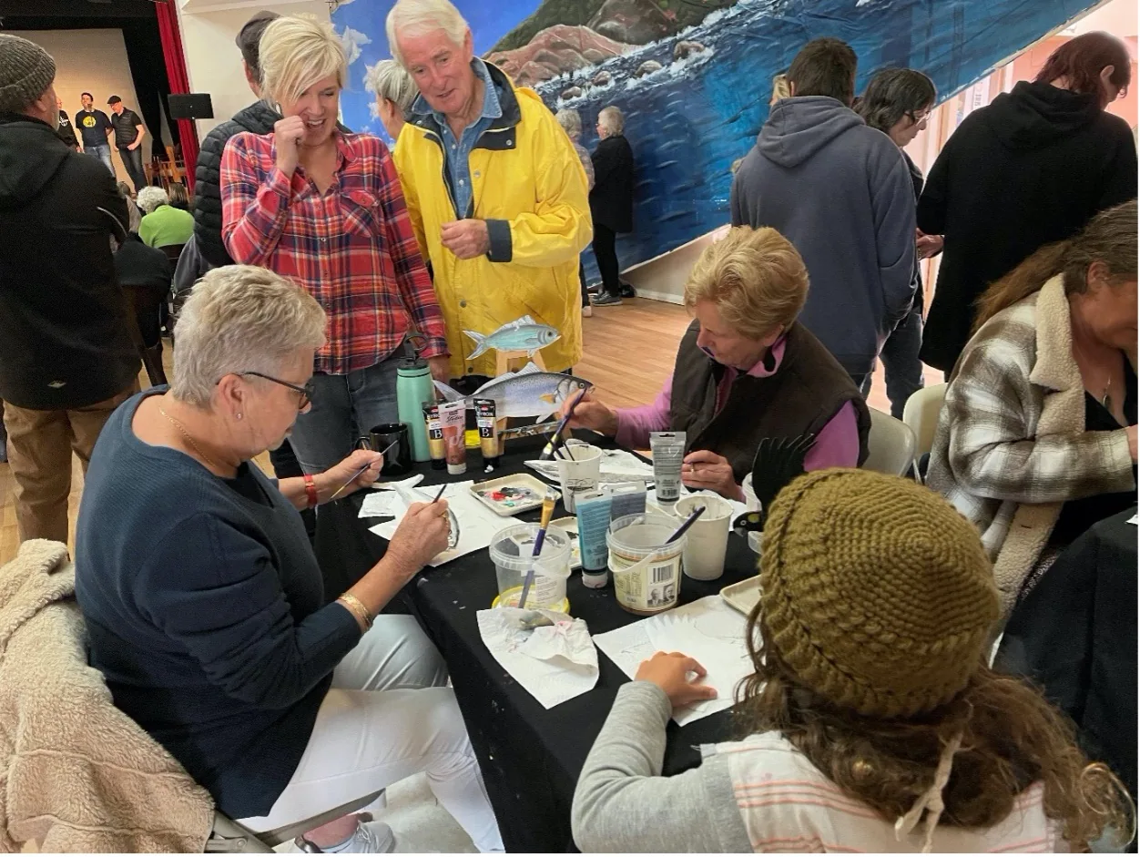 Volunteers painting small fish cutouts at tables in front of the sail mural