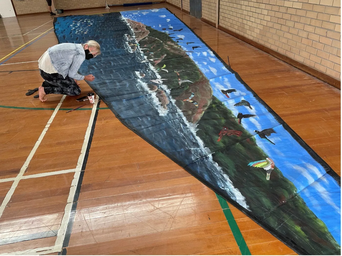 Artist kneeling to paint the coastal mural directly onto the laid-out sail