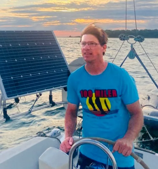 Man at the helm in a "100 Miler" shirt beside a solar panel at sunset