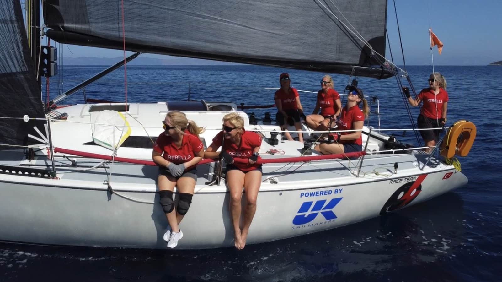 Team Rebella sailors in red shirts sit on the rail, 'Powered by UK' on the hull