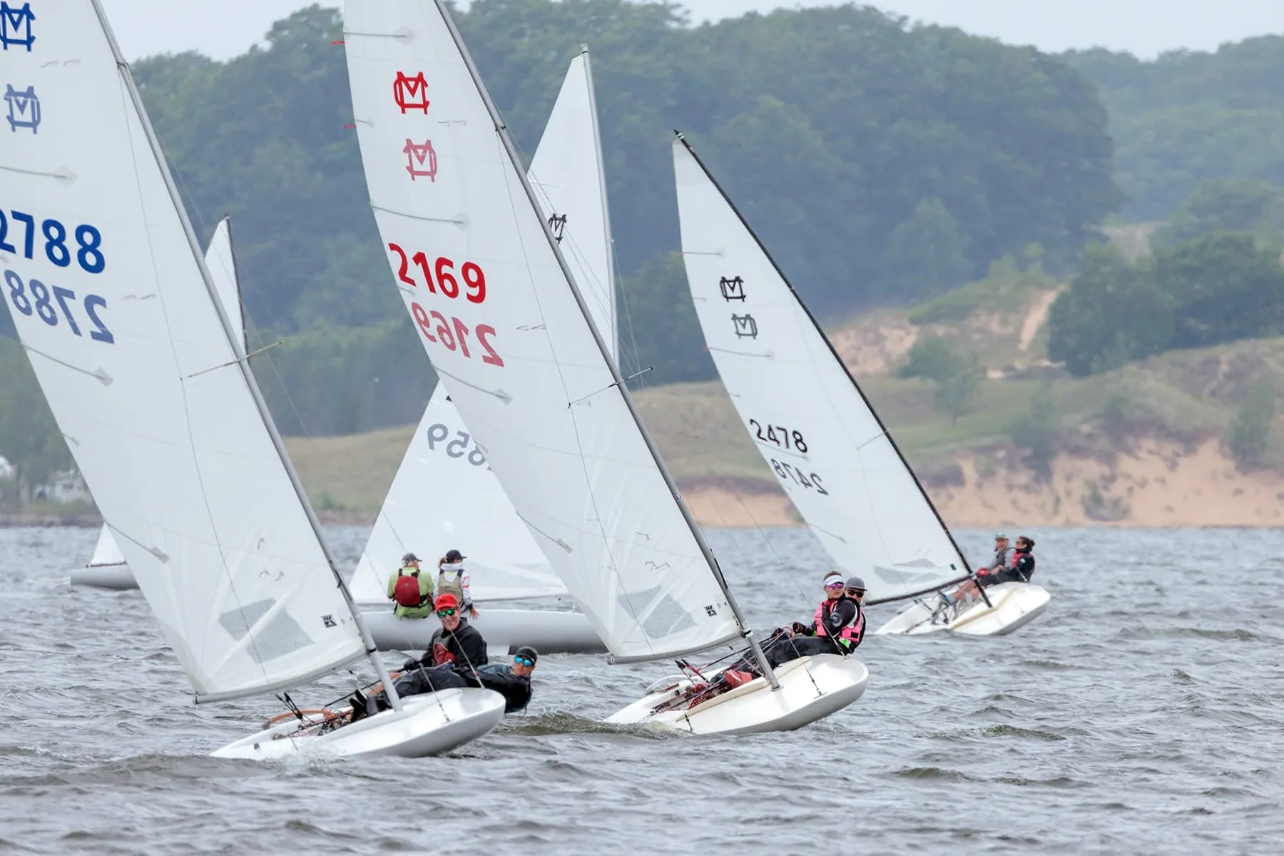 MC Scow fleet racing upwind on a lake with wooded shoreline