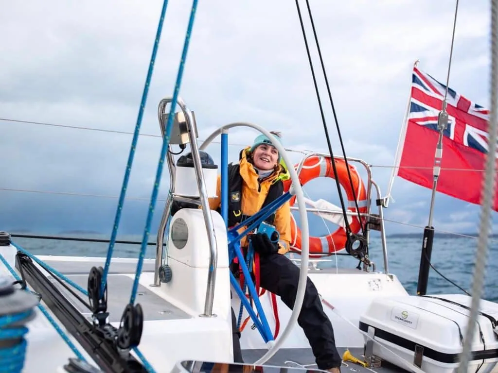 Sailor at the helm in foul-weather gear with a UK ensign flying astern