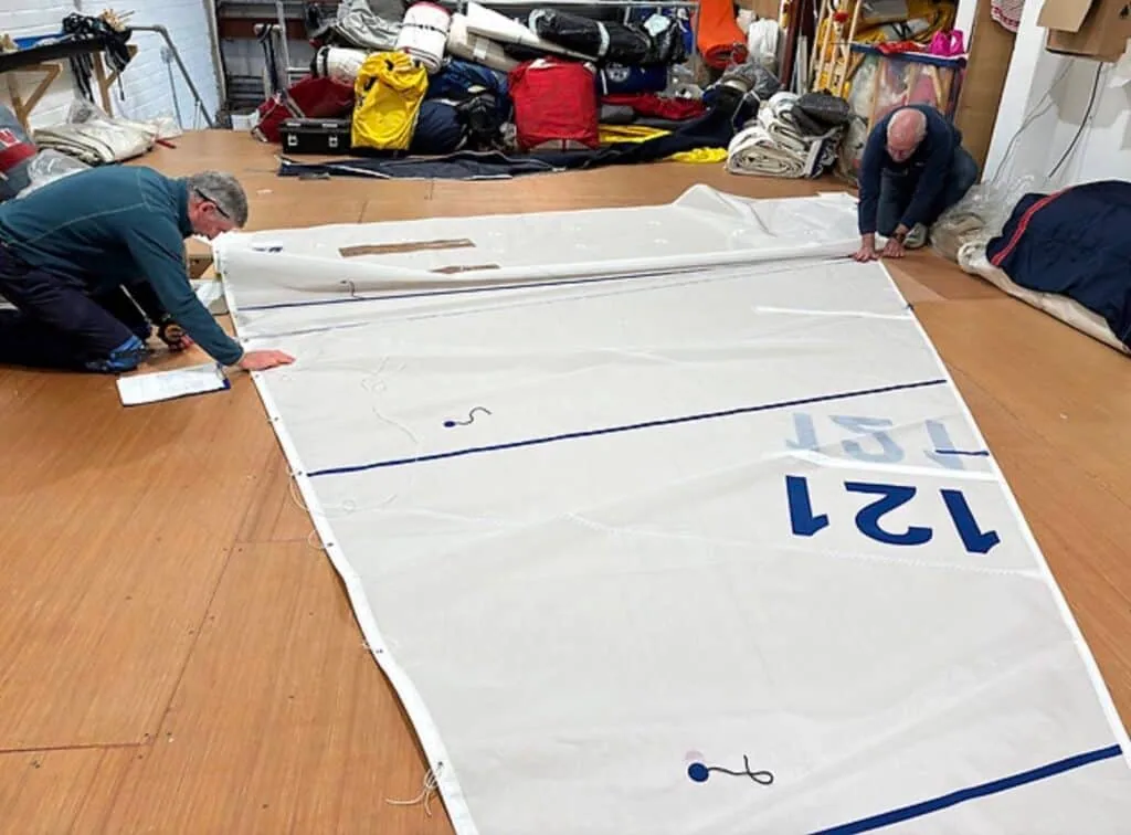 Two sailmakers rolling and marking a finished sail numbered 121 on the loft floor