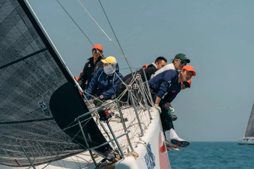 Crew hiking out on BIG APPLE III's bow with orange-and-white hull graphics