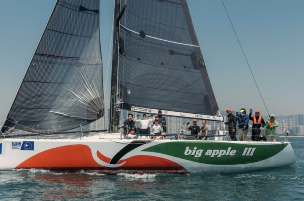 BIG APPLE III under black sails with her name visible on the orange-and-white hull