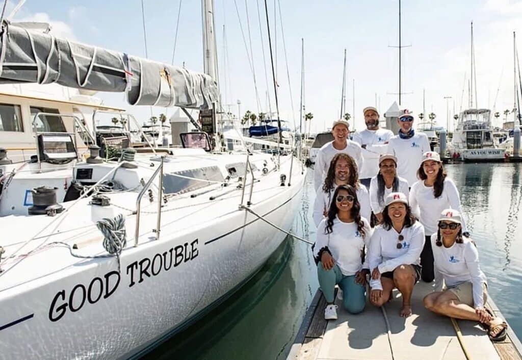 GOOD TROUBLE crew posing on the dock beside the sailboat's bow in a marina