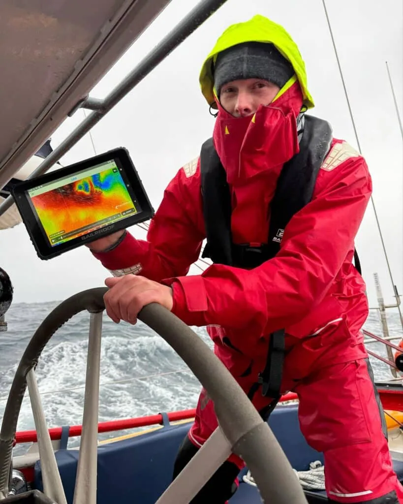 Sailor at the helm in foul-weather gear holding a tablet showing a sea-surface temperature map