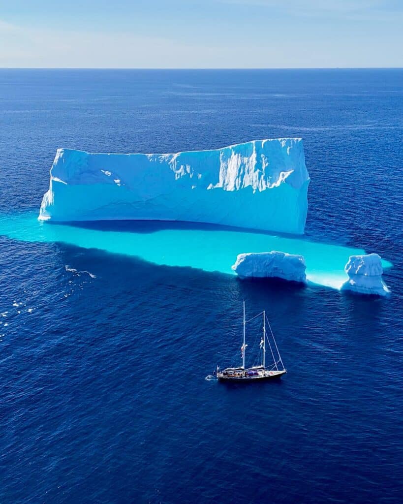 Two-masted sailboat anchored beside a large tabular iceberg with turquoise submerged ice