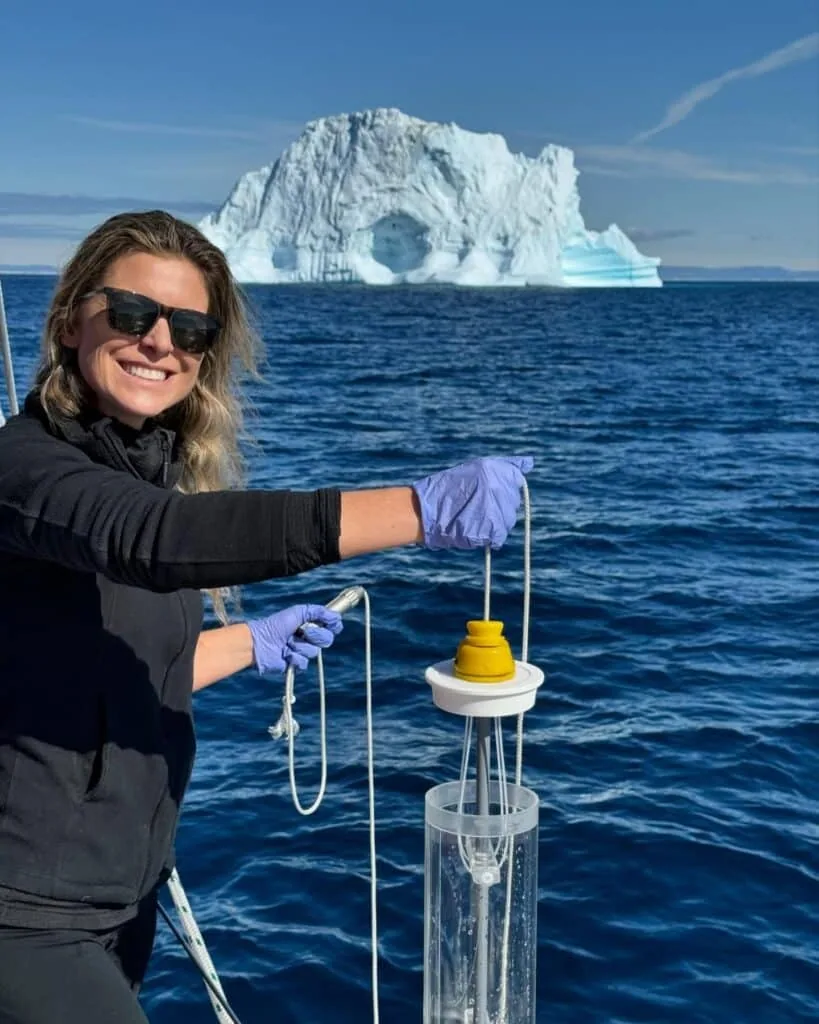 Scientist holding a water-sampling instrument on deck with an iceberg behind her