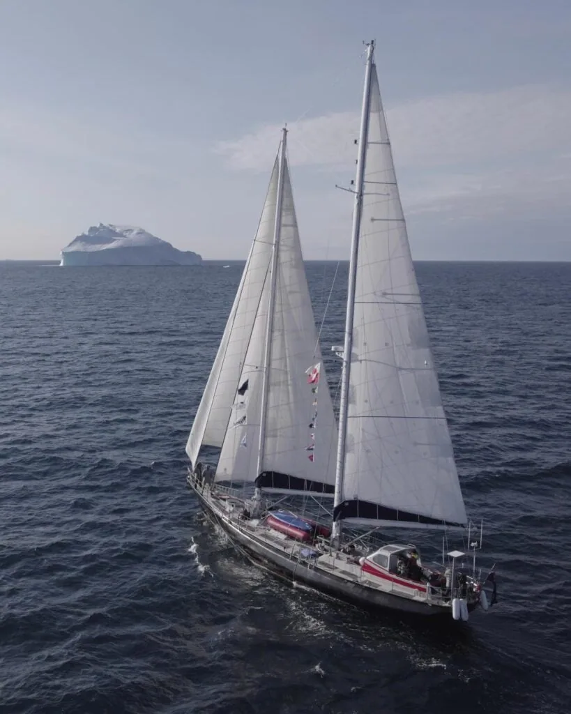 Two-masted sailboat under full sail with a distant iceberg on the horizon