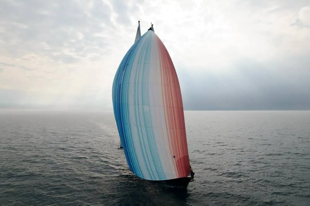 Aerial view of ABEL TASMAN's striped Climate Stripes gennaker filled with wind over open water.