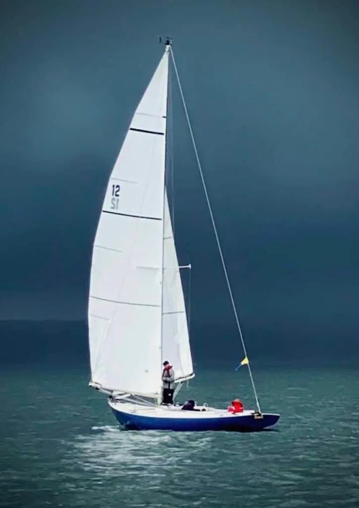 A solo sailor helms yacht 12 SI across calm water under a dark, stormy sky.