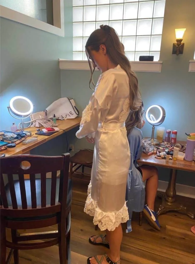 Model in a satin robe stands at a lit vanity mirror during a behind-the-scenes fitting.