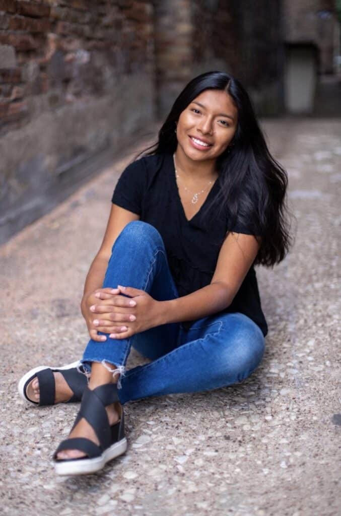 Portrait of a smiling young woman with long dark hair sitting on a stone walkway.