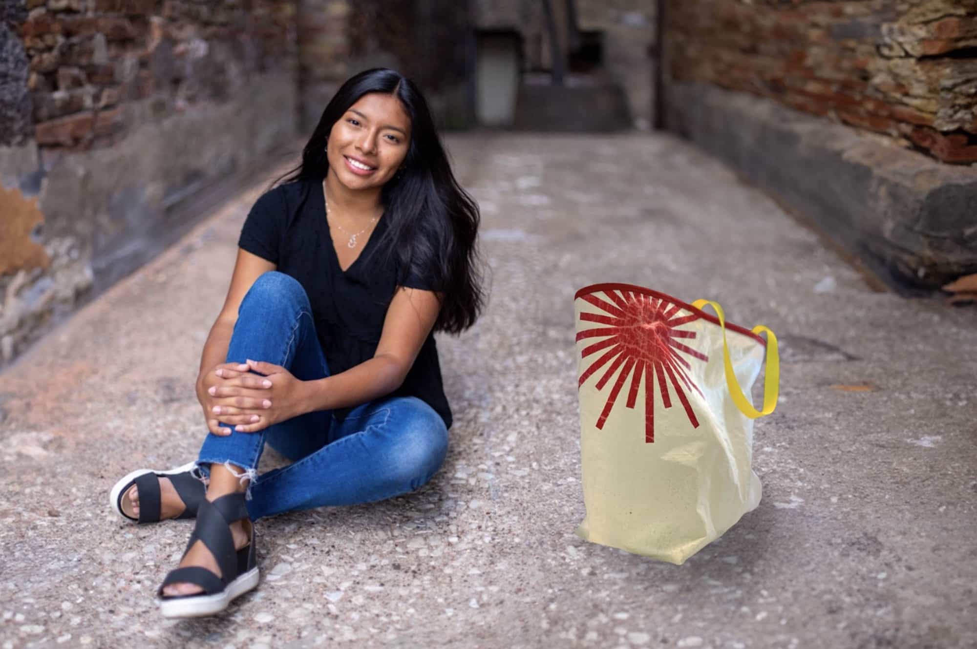 Young woman sits on a stone path beside a cream sailcloth tote with a red sunburst design.