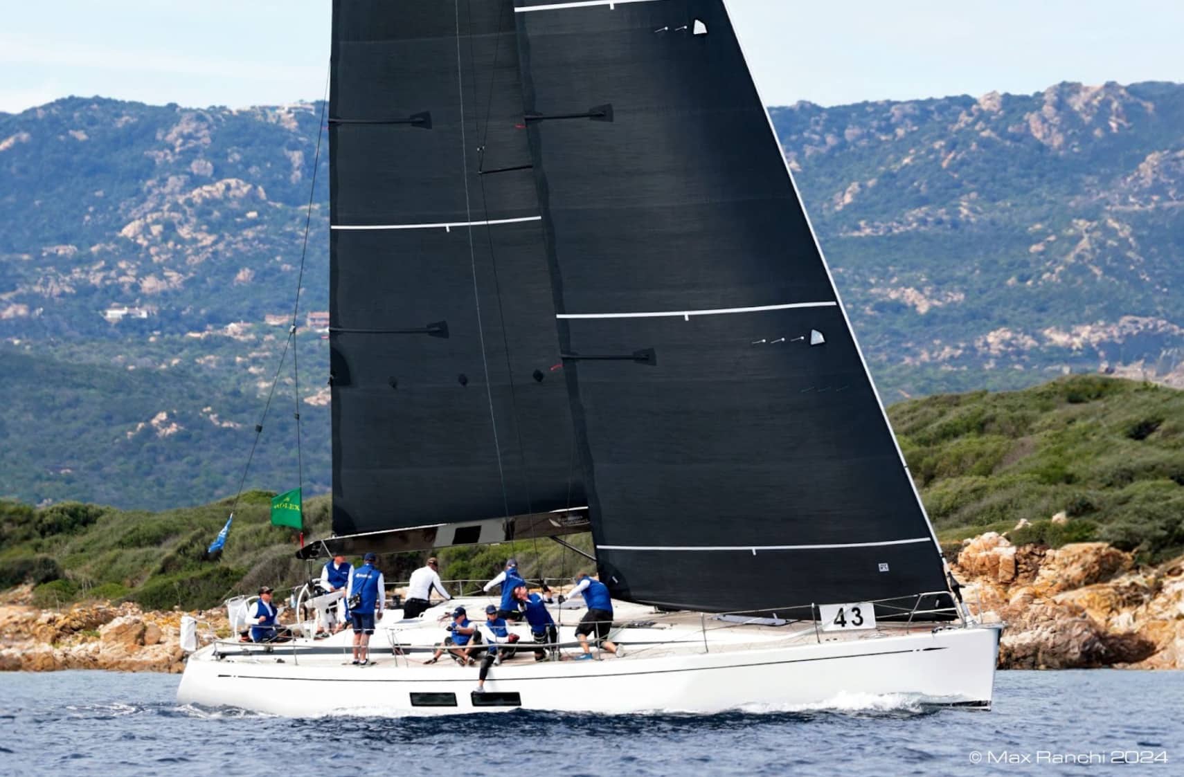 Yacht 43 sails hard under black sails off a rocky, hilly coastline at the Rolex Swan Cup.