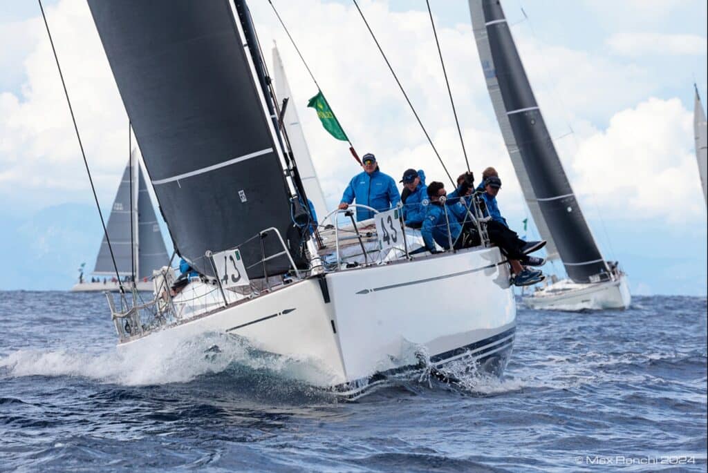 Swan yacht heels hard with crew hiked out under black sails, spray flying off the bow.