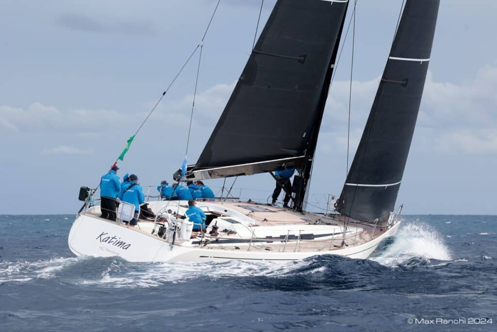 Swan 54 KATIMA heels under black sails with crew in blue hiked out, spray flying.
