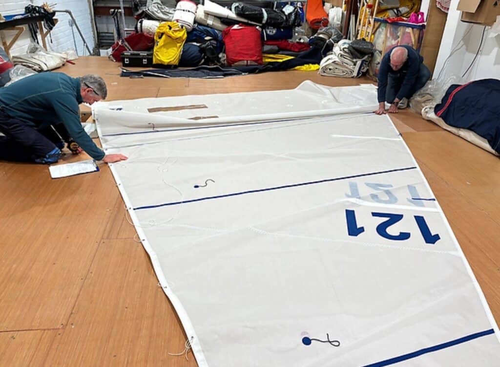 Two sailmakers inspect a numbered white sail laid out on the loft floor.