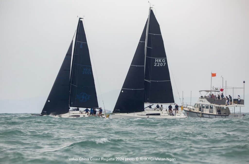 Two dark-sailed yachts, including HKG 2207, sail close together near a committee boat.