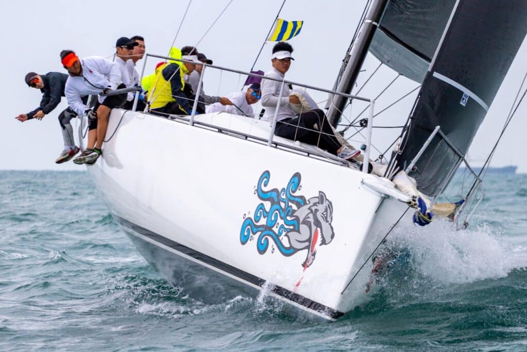 SEAWOLF heels hard with crew hiked out, her bow decorated with a cartoon wolf logo.