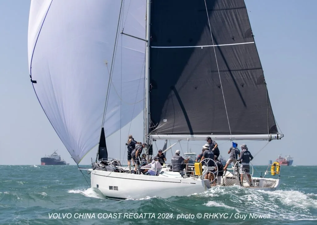 Yacht sails under a white asymmetric spinnaker and black mainsail, cargo ships astern.
