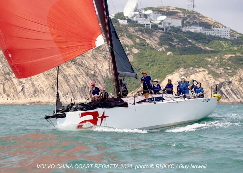 Yacht with a red star logo races under orange spinnaker past Hong Kong's radar-topped hills.