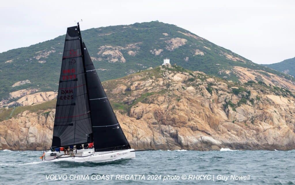 A yacht sails close-hauled under black sails past a rocky, green Hong Kong headland.