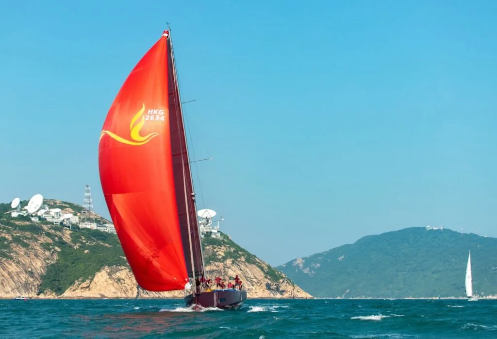 FENICE reaches under a red spinnaker with a gold flame logo past Hong Kong's hills.