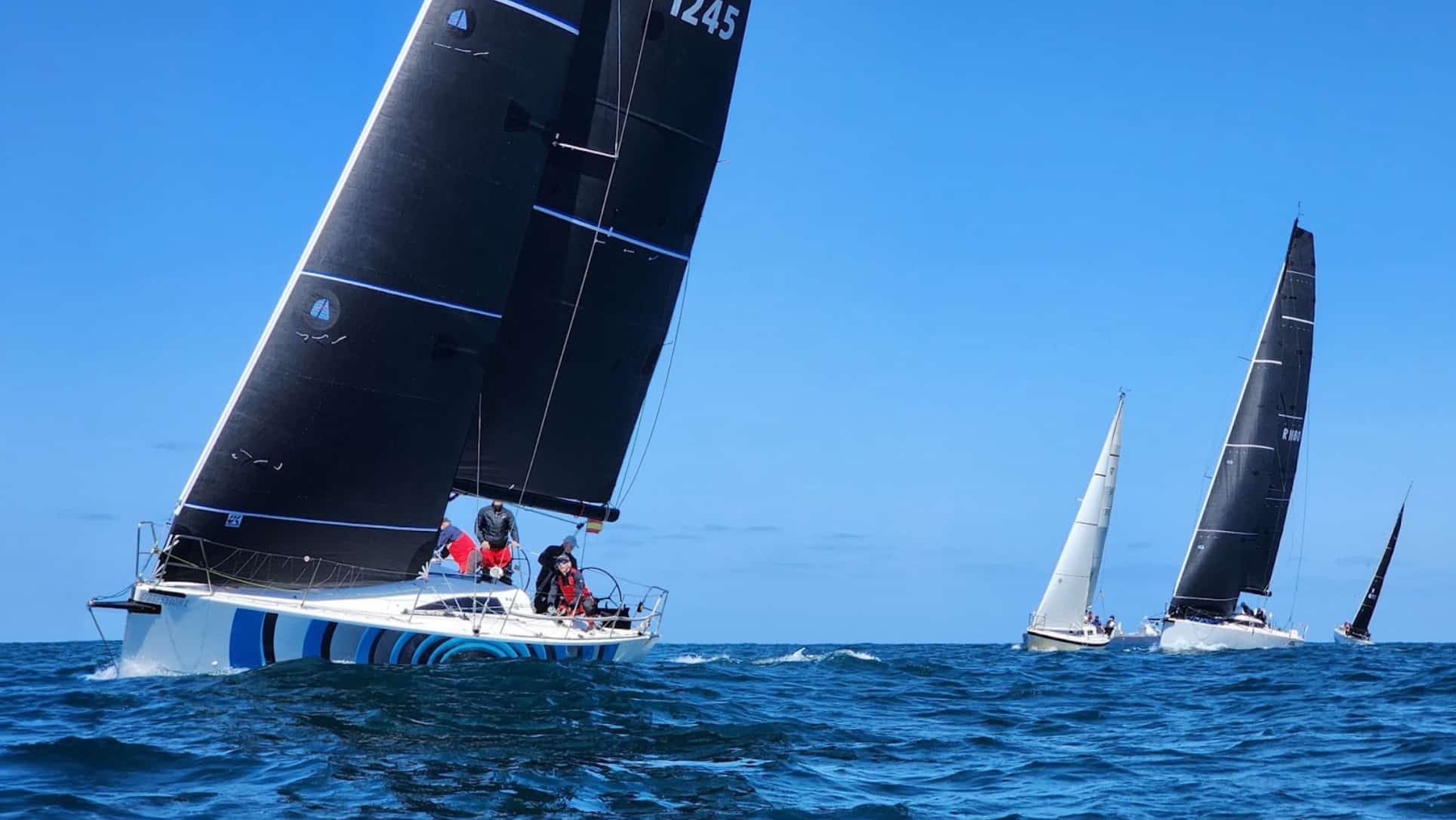 OBSESSION, hull 1245, sails under a black-striped mainsail with two yachts racing behind.