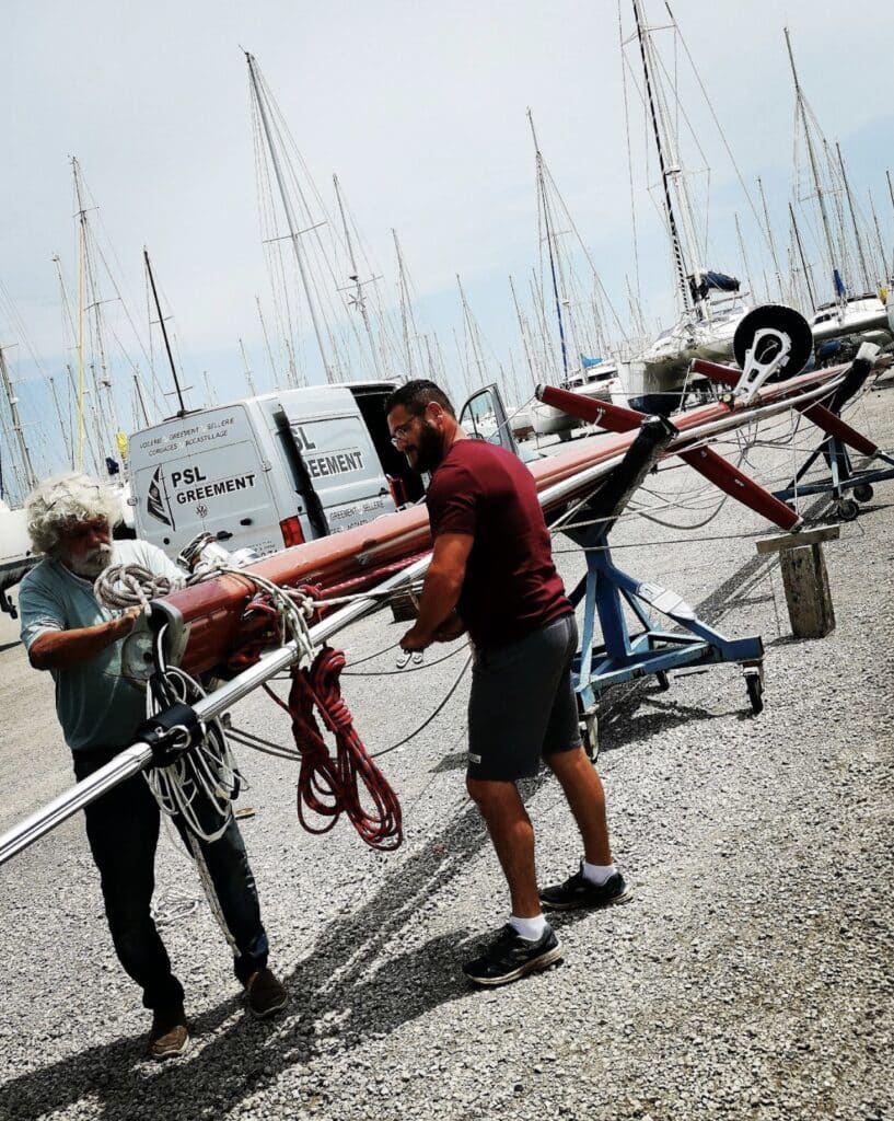 Two riggers carrying a spinnaker pole through a boatyard