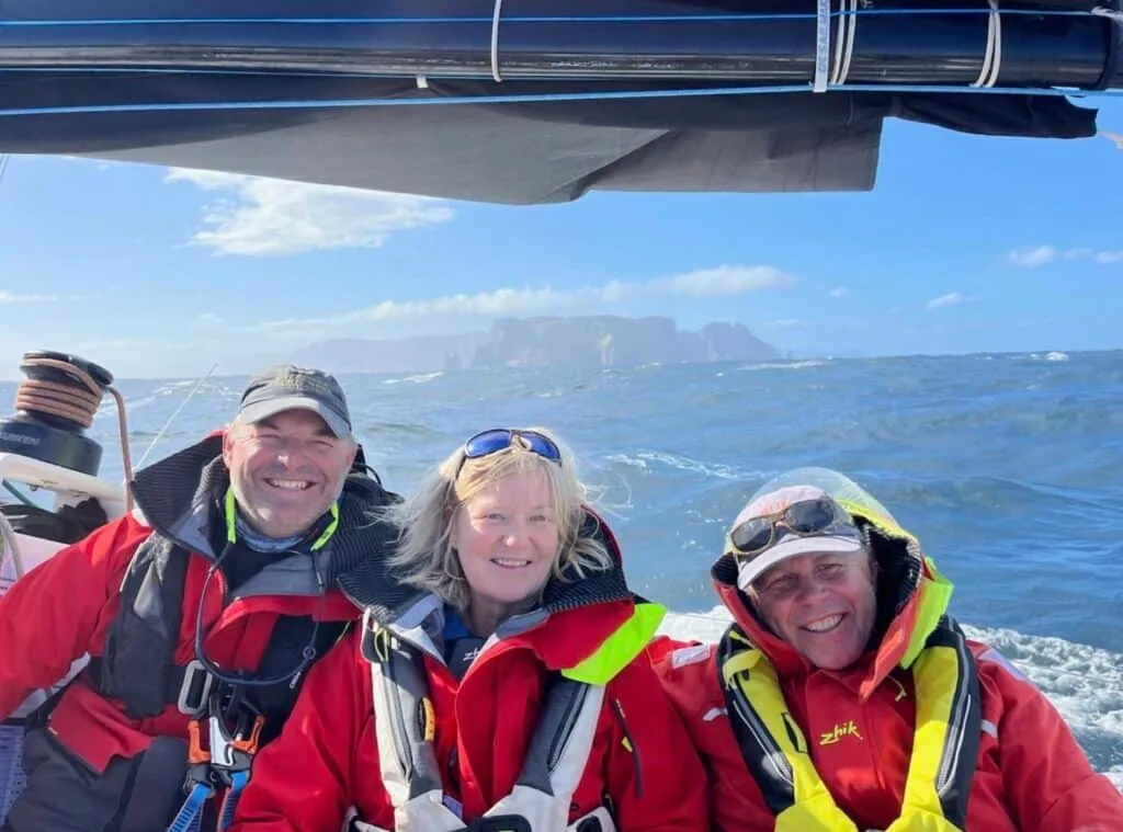 Three crew smiling on deck with Tasman Island's cliffs behind