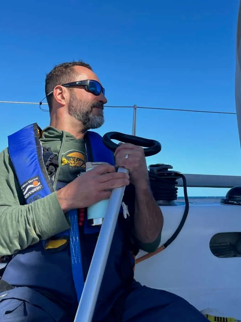Sailor at the wheel of SOULMATES wearing a life jacket, blue sky behind