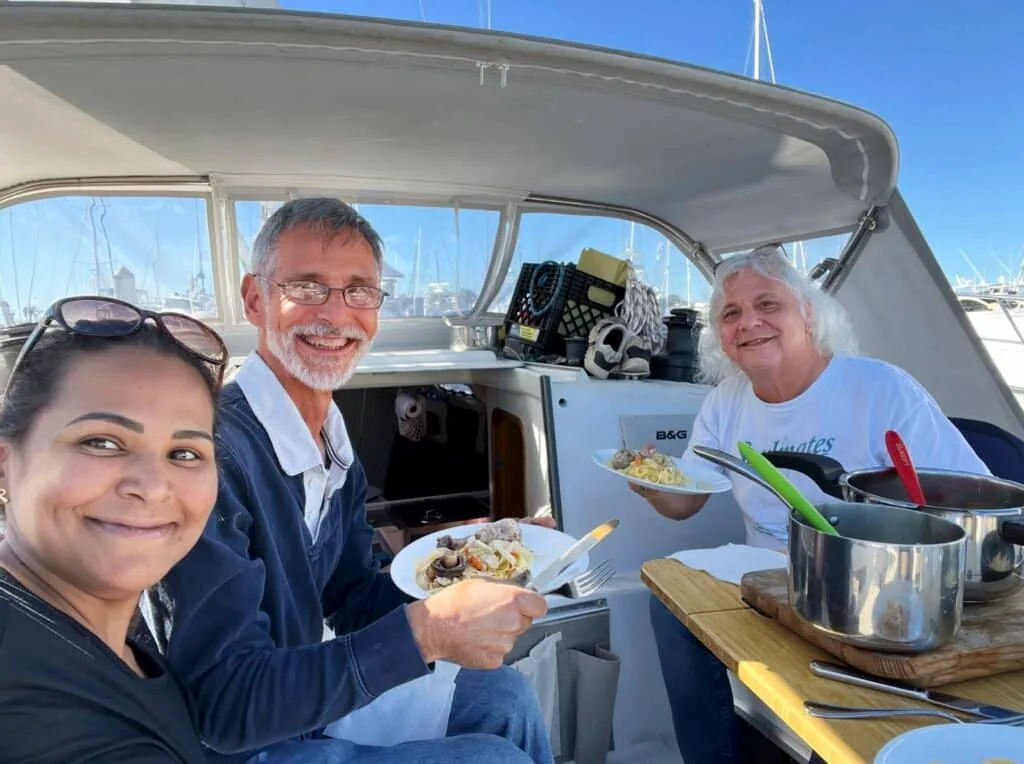 Three people sharing a meal in a sailboat's cockpit at a sunny marina