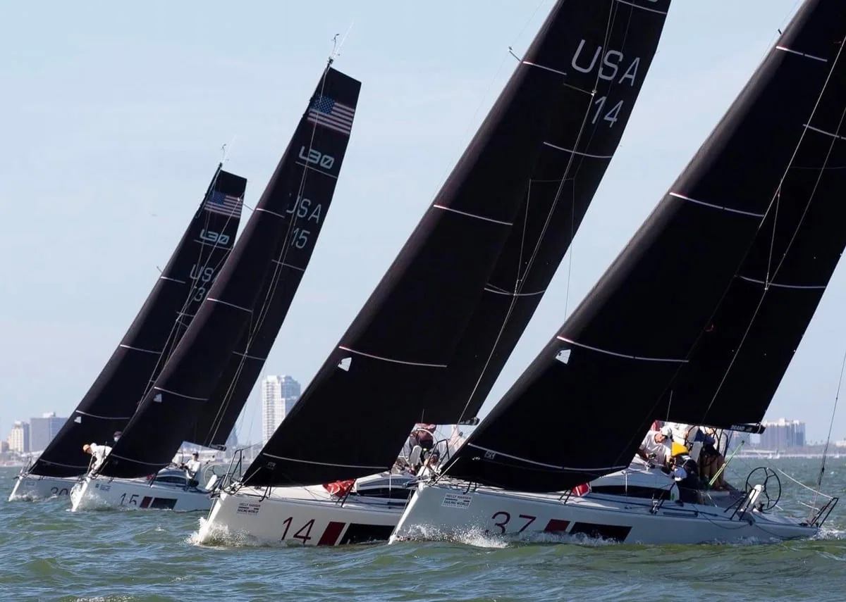 Black mainsails with bold white USA sail numbers on a fleet racing off a city skyline