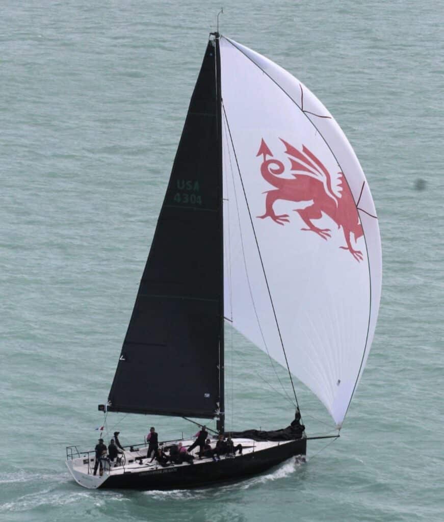 Yacht flying a white spinnaker with a red dragon emblem beside dark numbered sails