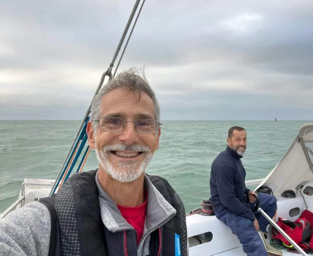 Selfie of a sailor at the helm with a crewmate seated forward, cloudy sky offshore