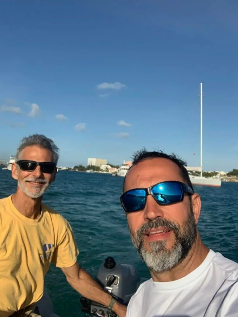 Two men taking a selfie on a boat with a calm harbor and buildings behind them