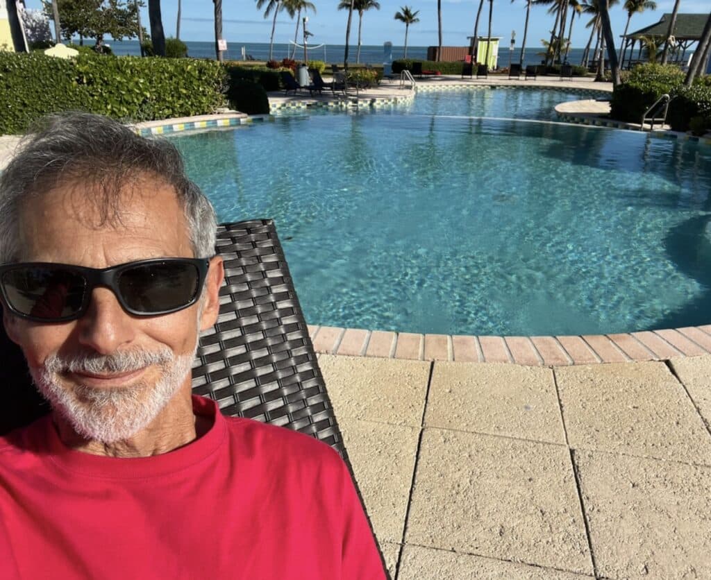 Man in a red shirt and sunglasses sitting poolside near palm trees and the ocean