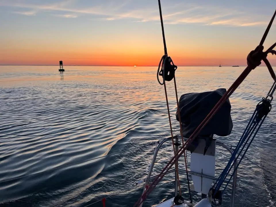 Sunset over calm water from a sailboat's bow, with a channel buoy ahead