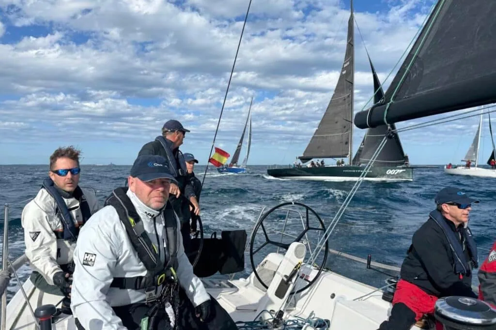 Crew at the helm on a breezy day, racing fleet including a Spanish-flagged boat astern