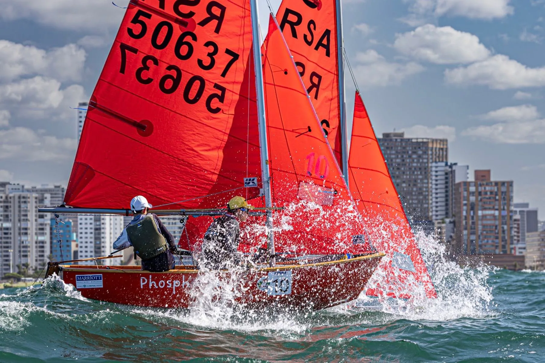 Mirror dinghies with red sails crest a wave, Durban skyline behind them